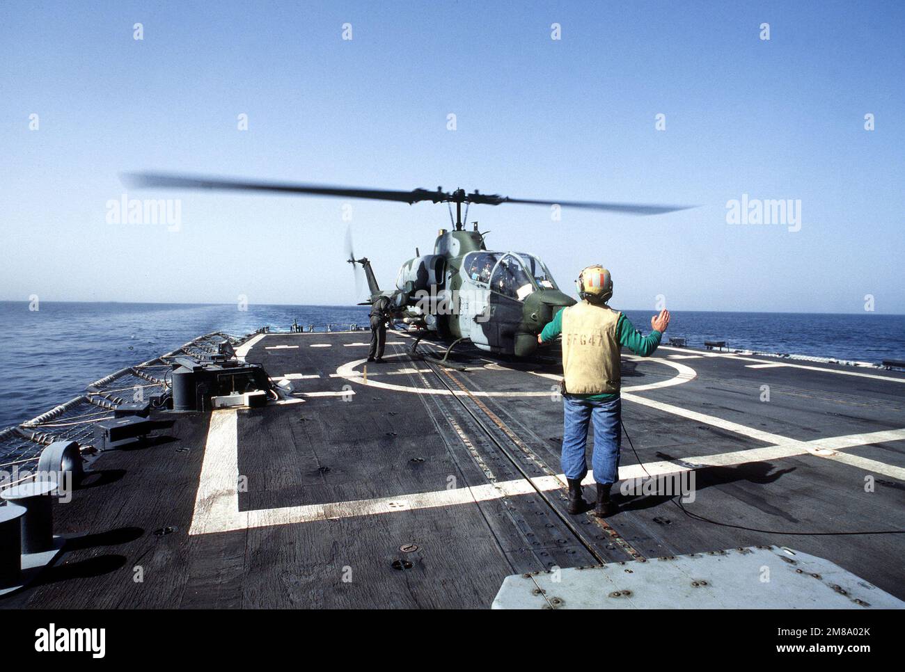 Un direttore dell'aereo segnala al pilota di una nave da guerra Marine Utility/Attack Helicopter Squadron 257 (HMLA-257) AH-1 Sea Cobra sul ponte dell'elicottero della fregata missilistica guidata USS NICHOLAS (FFG-47) durante la volontà seria, Una missione di convoglio in cui le petroliere Kuwaitane riflesse sono scortate attraverso le acque del Golfo da navi navali statunitensi. Oggetto operazione/Serie: SERNEST WILL Paese: Sconosciuto Foto Stock
