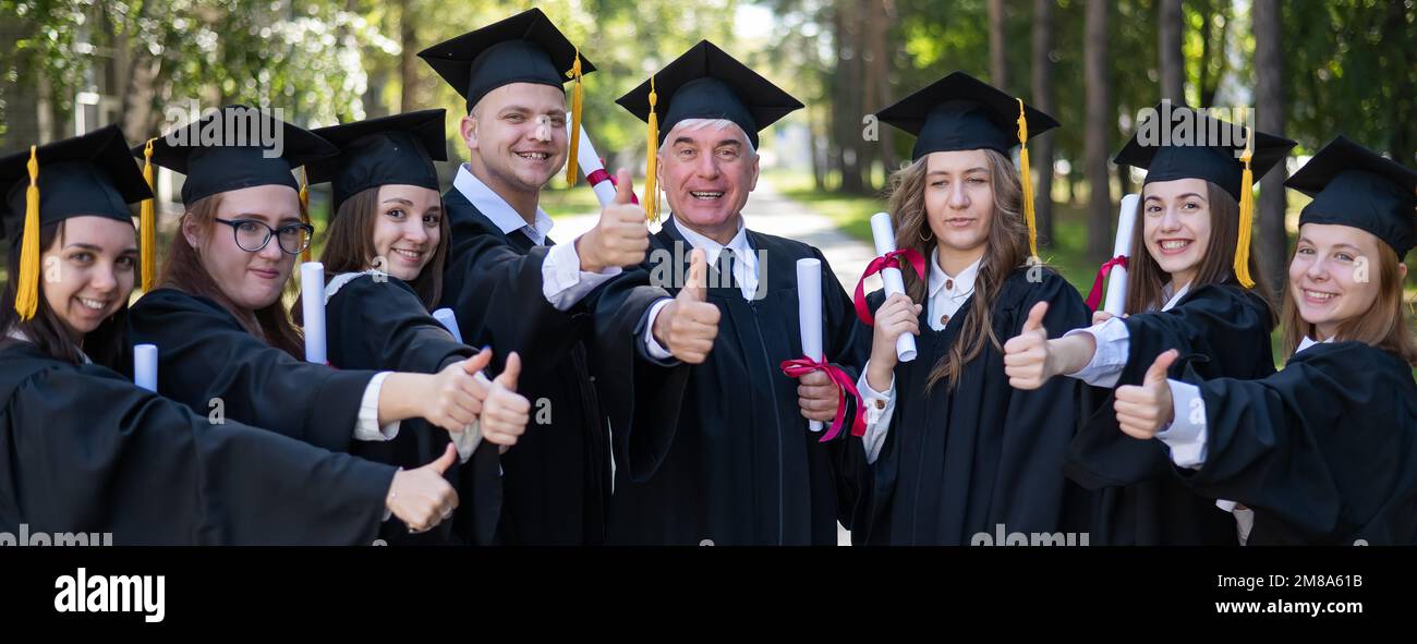 Un gruppo di laureati in vesti danno un pollice in su all'aperto. Studente anziano. Foto Stock