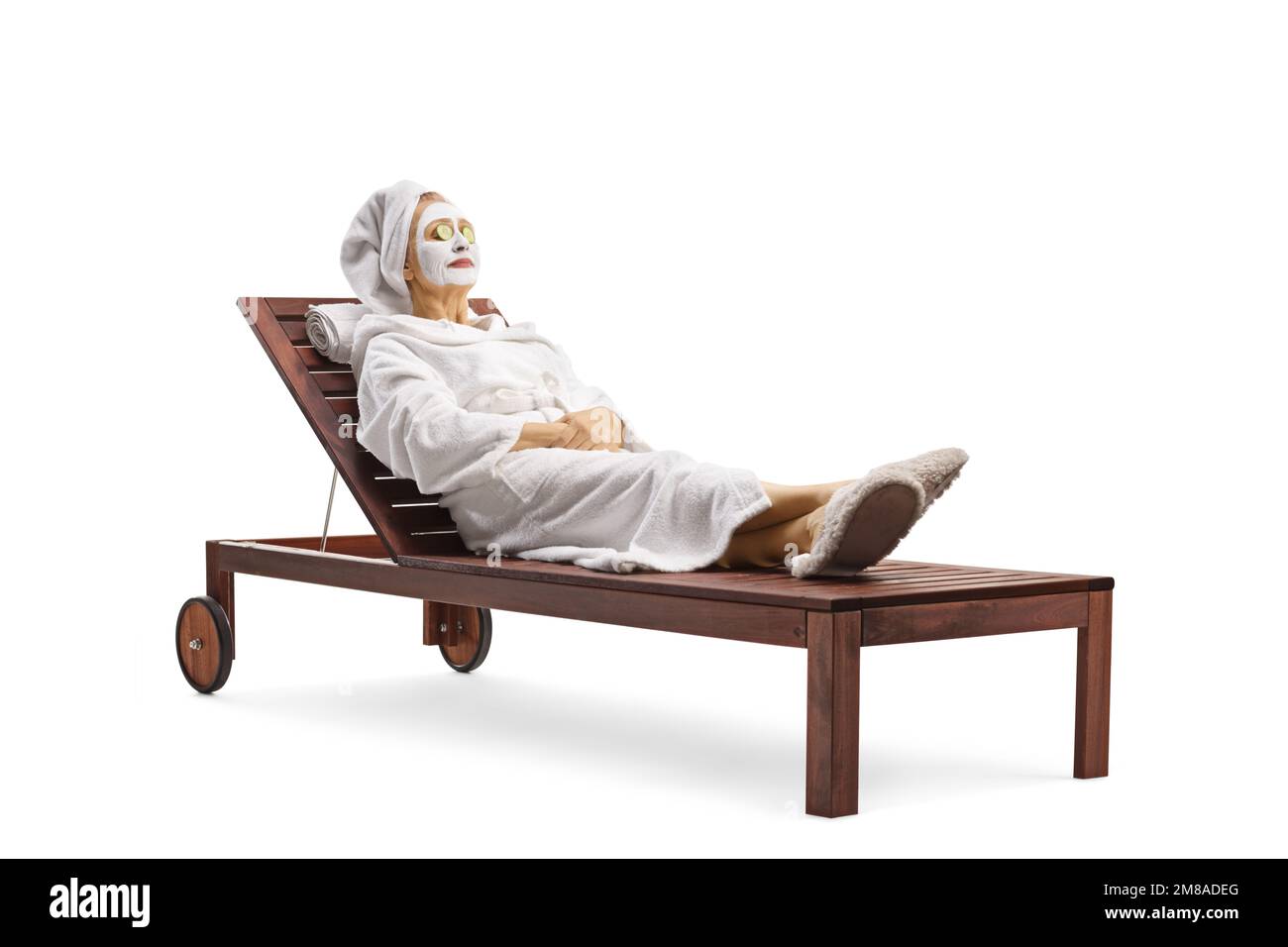 Donna matura in un accappatoio e capelli avvolti in un asciugamano posando con una maschera facciale e cetrioli sugli occhi isolati su sfondo bianco Foto Stock