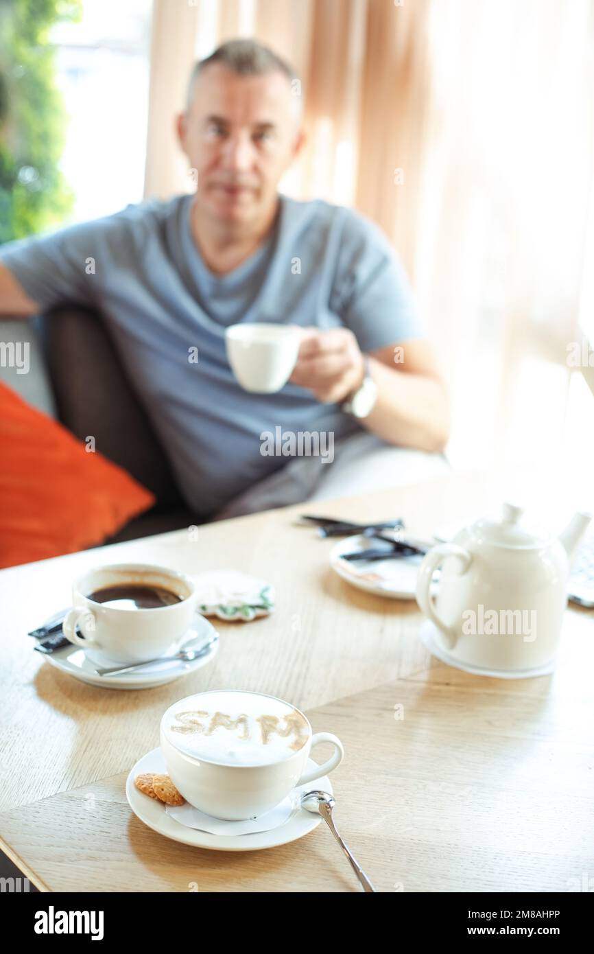 Tazza da caffè verticale, crema segno schiuma SMM di cannella con biscotti, teiera sul tavolo da caffè. Un uomo d'affari offuscato beve il tè Foto Stock