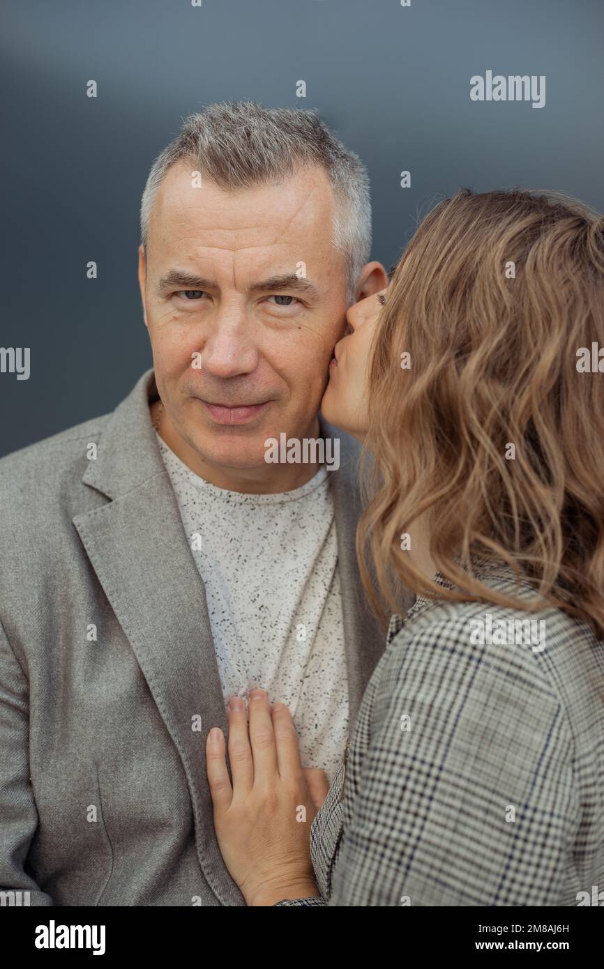 Coppia verticale romantica di due mature uomo e giovane donna dai capelli grigi che baciano alla guancia, stropicciando su sfondo grigio Foto Stock