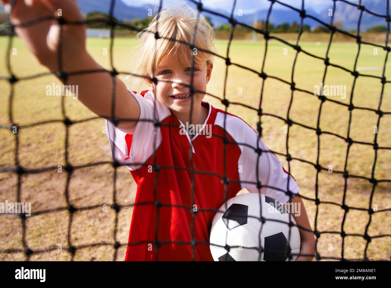 Il suo obiettivo è in vista. Una bambina che tiene una sfera di calcio mentre guarda attraverso la rete di obiettivo su un campo di calcio. Foto Stock