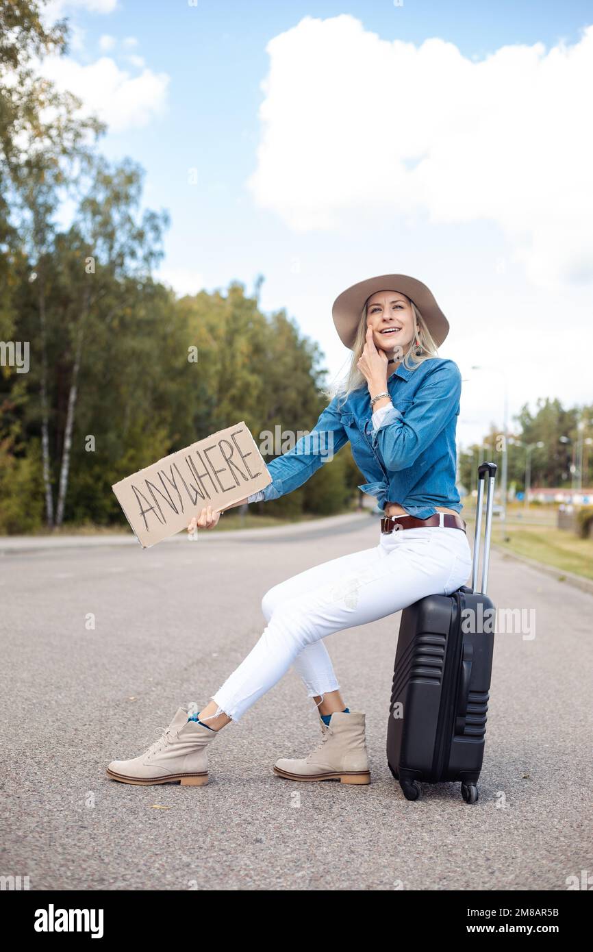 Donna bionda sorridente e ridente verticale in cowgirl cappello hitchhike da strada con piastra di cartone ovunque sedersi sulla valigia Foto Stock