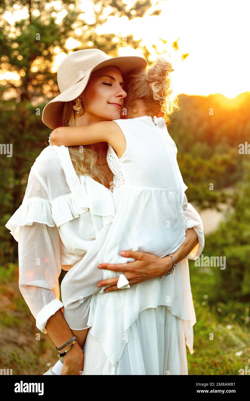 Ritratto di felice donna sorridente in cappello con amore tenere in mano bambina all'aperto in legno al tramonto. Mamma e bambino a piedi nella foresta in estate Foto Stock