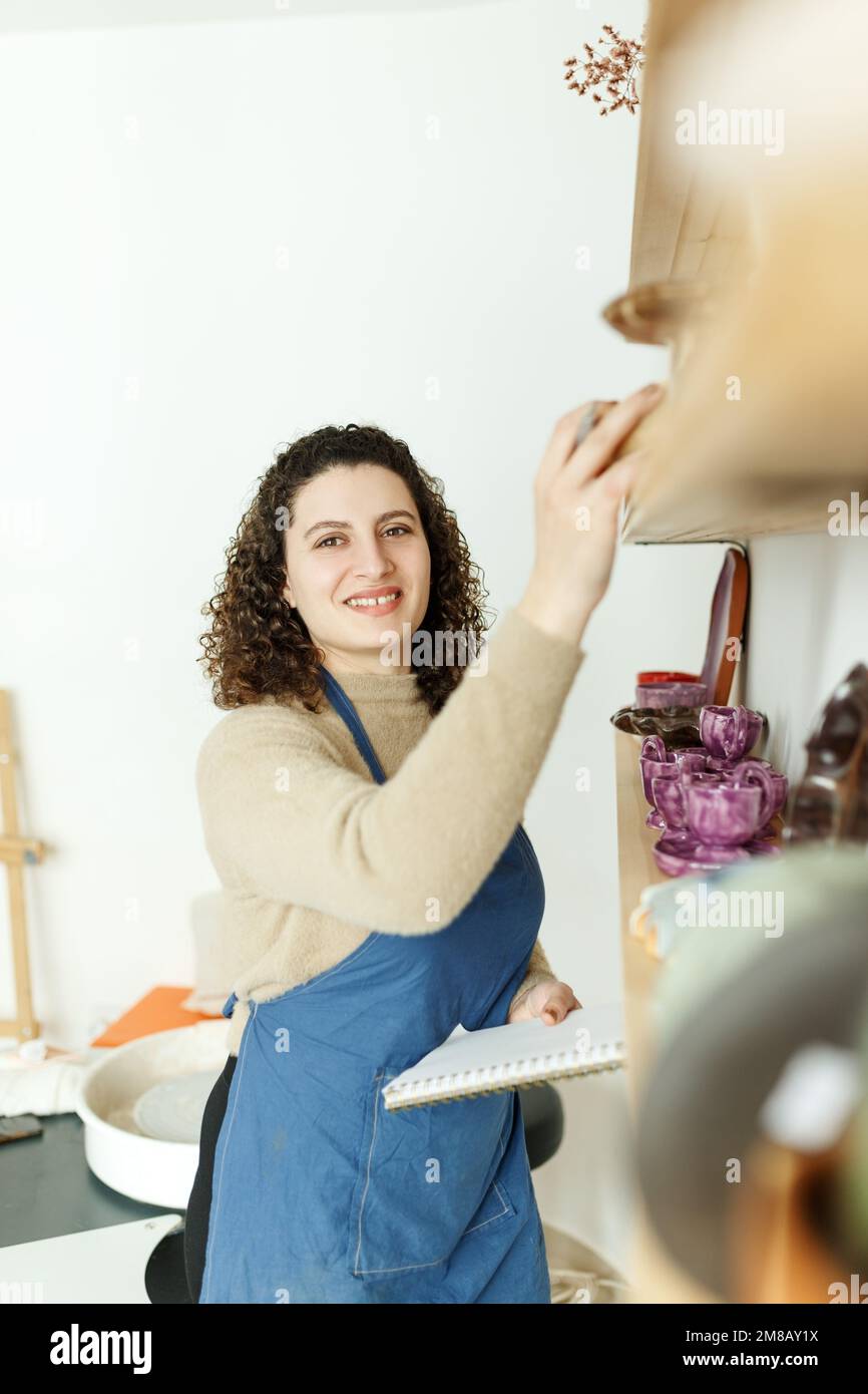Felice giovane ceramista guardando via pensieroso mentre prende il calcio. Donna d'affari creativa che gestisce un negozio con prodotti in ceramica fatti a mano. Interno femminile Foto Stock