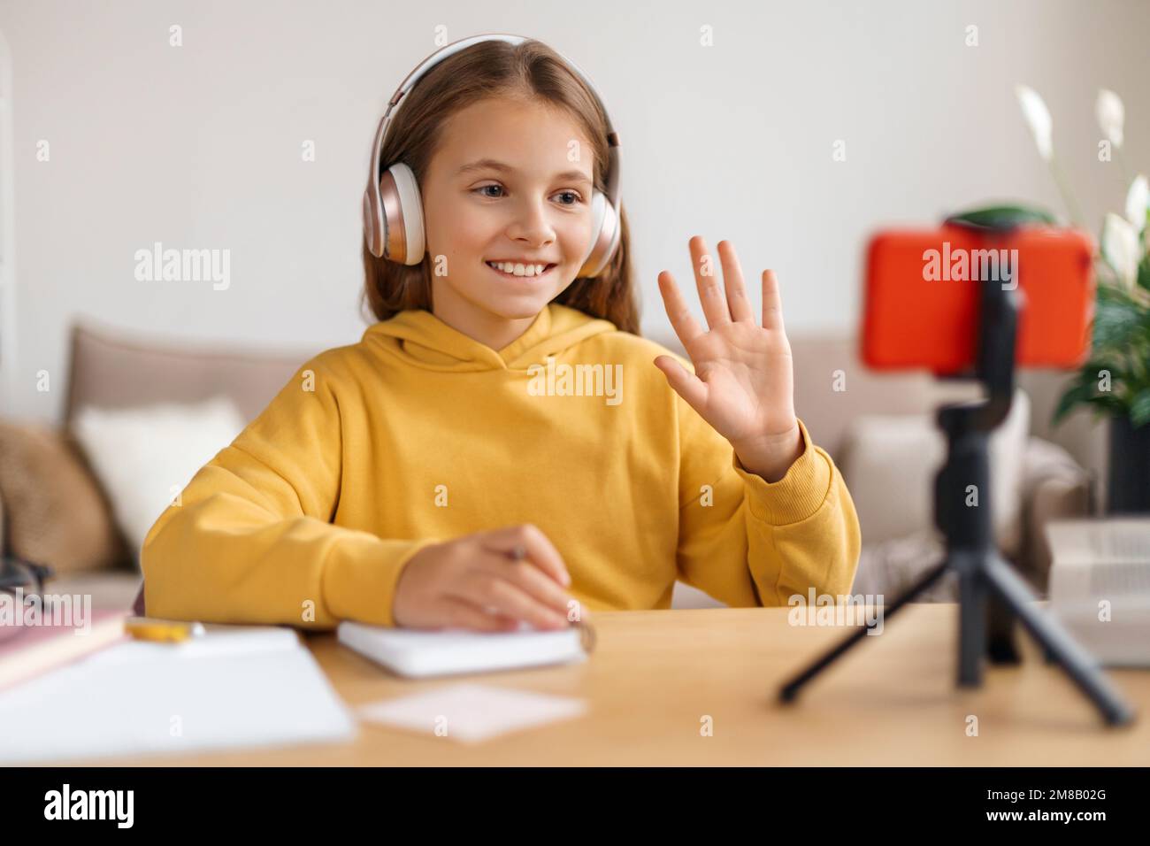 Ragazza della scuola positiva che ha una videoconferenza sullo smartphone e una lezione online con un insegnante da casa, indossando cuffie, sorridendo, muovendo la mano sullo schermo. Homeschooling, istruzione remota durante la quarantena Foto Stock