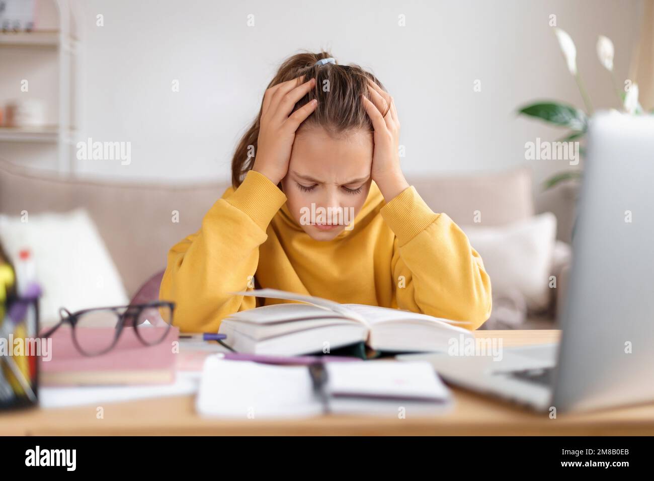 Ragazza scolastica esausta che soffre di fare i compiti a casa, stanca di studiare a casa, preparandosi prima degli esami, stressata con un compito difficile, leggendo un libro, utilizzando il computer portatile. Educazione domestica, homeschooling Foto Stock