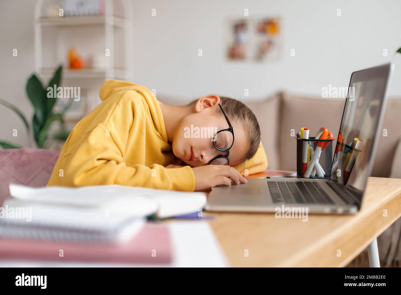Stanca bambina scolastica in bicchieri che dormiva alla scrivania di fronte al computer portatile a casa, si stanca di studiare, si addormentava mentre facevo i compiti a casa, guardava una lezione online o cercava informazioni su Internet Foto Stock