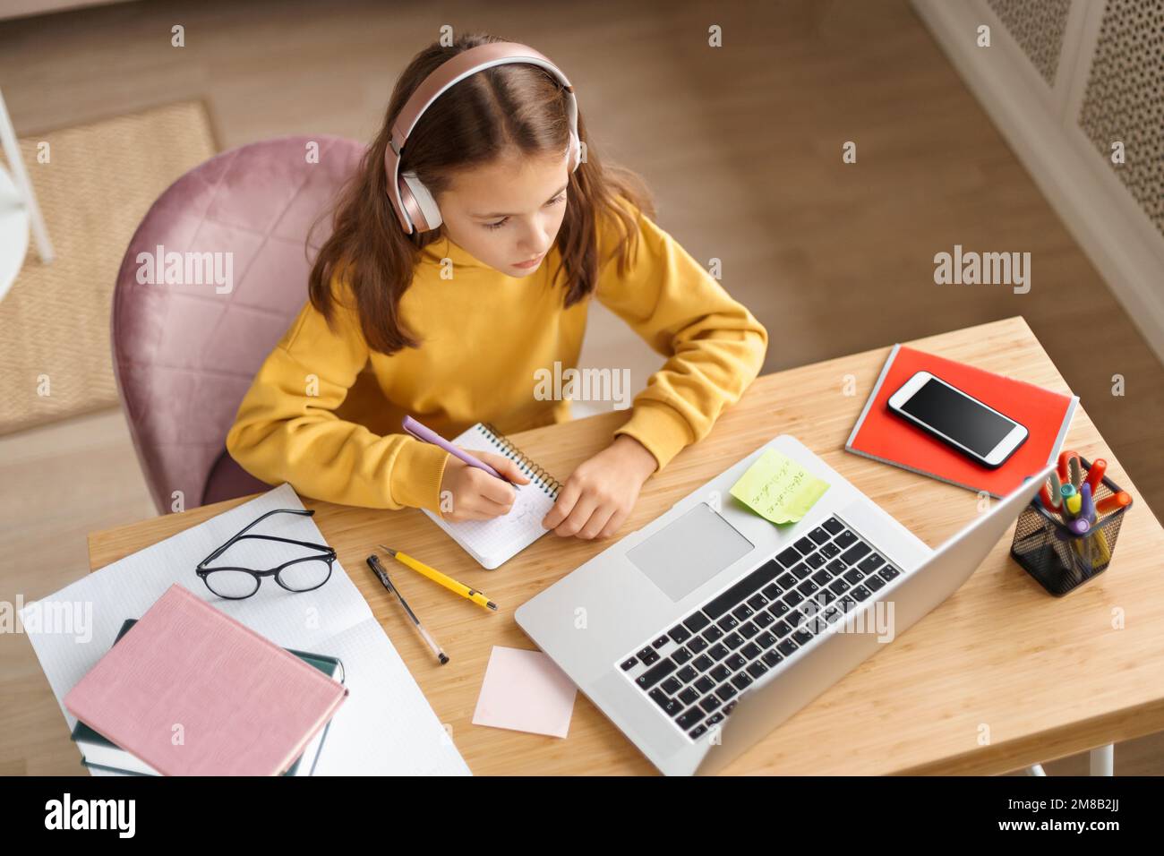 Vista dall'alto della giovane ragazza che indossa cuffie che fanno i compiti a casa sulla scrivania della sua camera, che usa un computer portatile, Internet, che ha una lezione online con un insegnante a distanza. Homeschooling durante la quarantena di covid Foto Stock