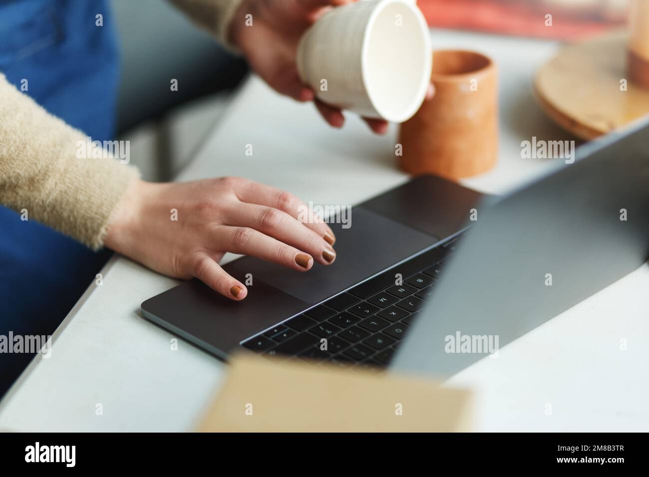 Potter imprenditore utilizzando il laptop in officina, solo le mani, tenendo in mano la tazza di ceramica, prende ordini online. Foto Stock