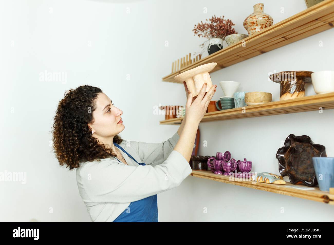 Ragazza giovane donna d'affari nel suo laboratorio è in piedi accanto a scaffale con prodotti in ceramica. Concetto di start-up per piccole imprese. Foto Stock