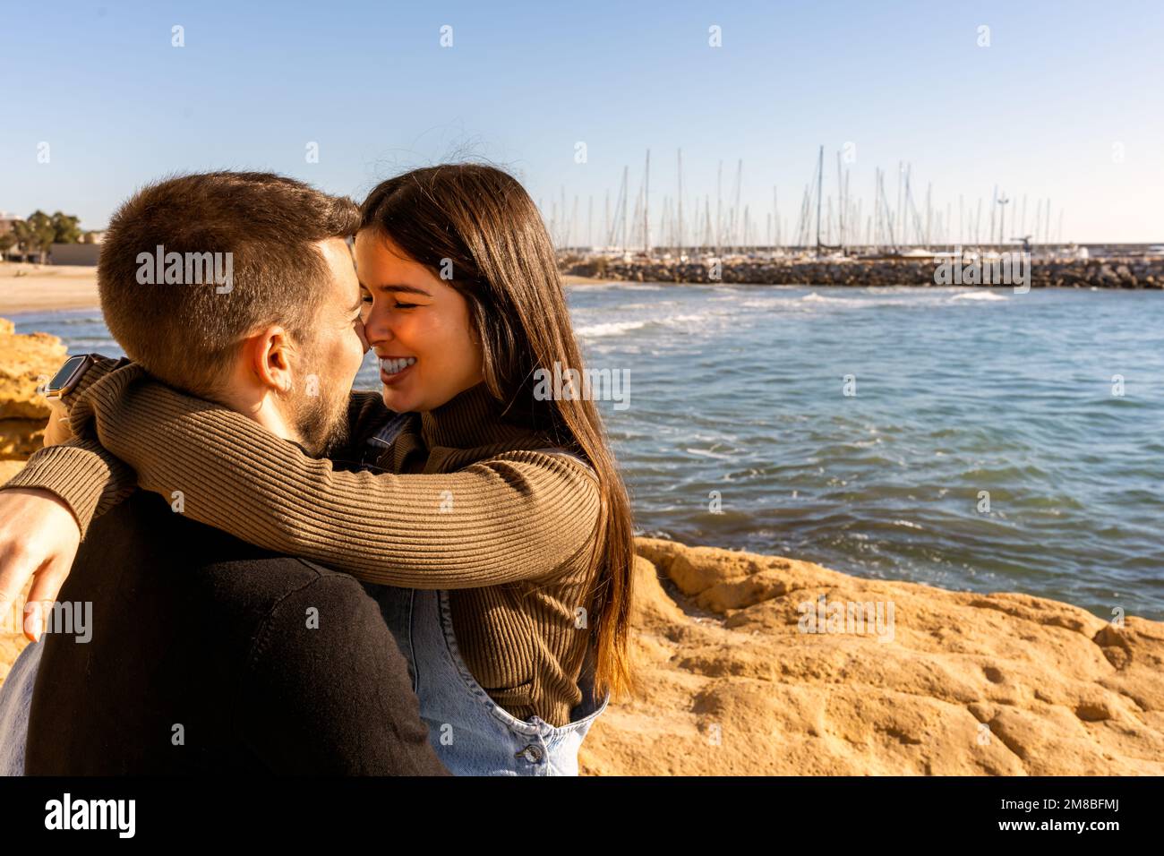 Giovane donna ottimista in abiti casual abbracciando e toccando nasi con l'uomo sulla spiaggia rocciosa il giorno di San Valentino Foto Stock
