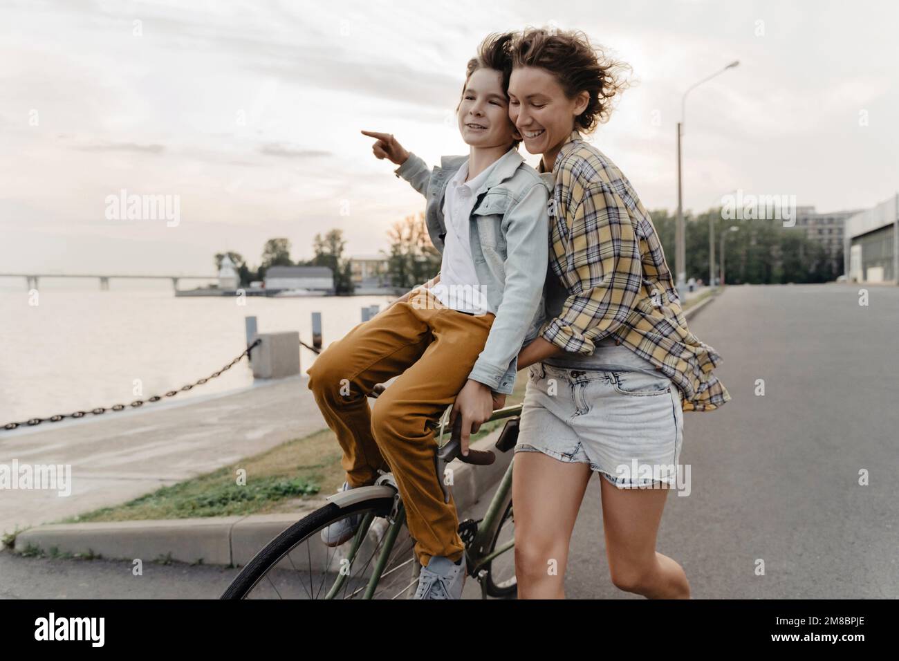 Giovane madre e figlio che si divertono mentre cavalcano in bicicletta. Ragazzino seduto sul manubrio di una bicicletta guidata dalla madre all'aperto. Foto Stock