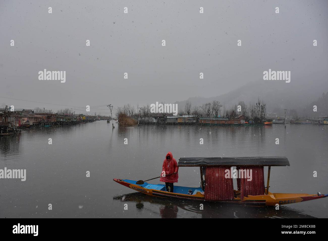 Un uomo che indossa un impermeabile rema la sua barca sul lago dal durante le nevicate. Parti della valle di Kashmir compreso Srinagar ha ricevuto nevicate che portano alla chiusura della vitale Srinagar-Jammu autostrada nazionale e la cancellazione delle operazioni di volo, funzionari qui ha detto. Foto Stock