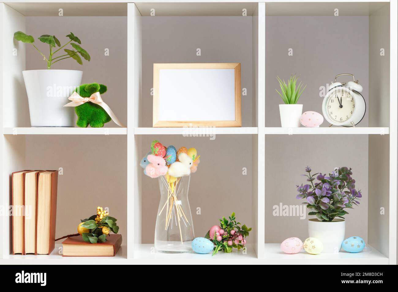 Interni per la casa, elementi decorativi pasquali. Mensole bianche con libri sveglia piante in vaso conigli uova Foto Stock