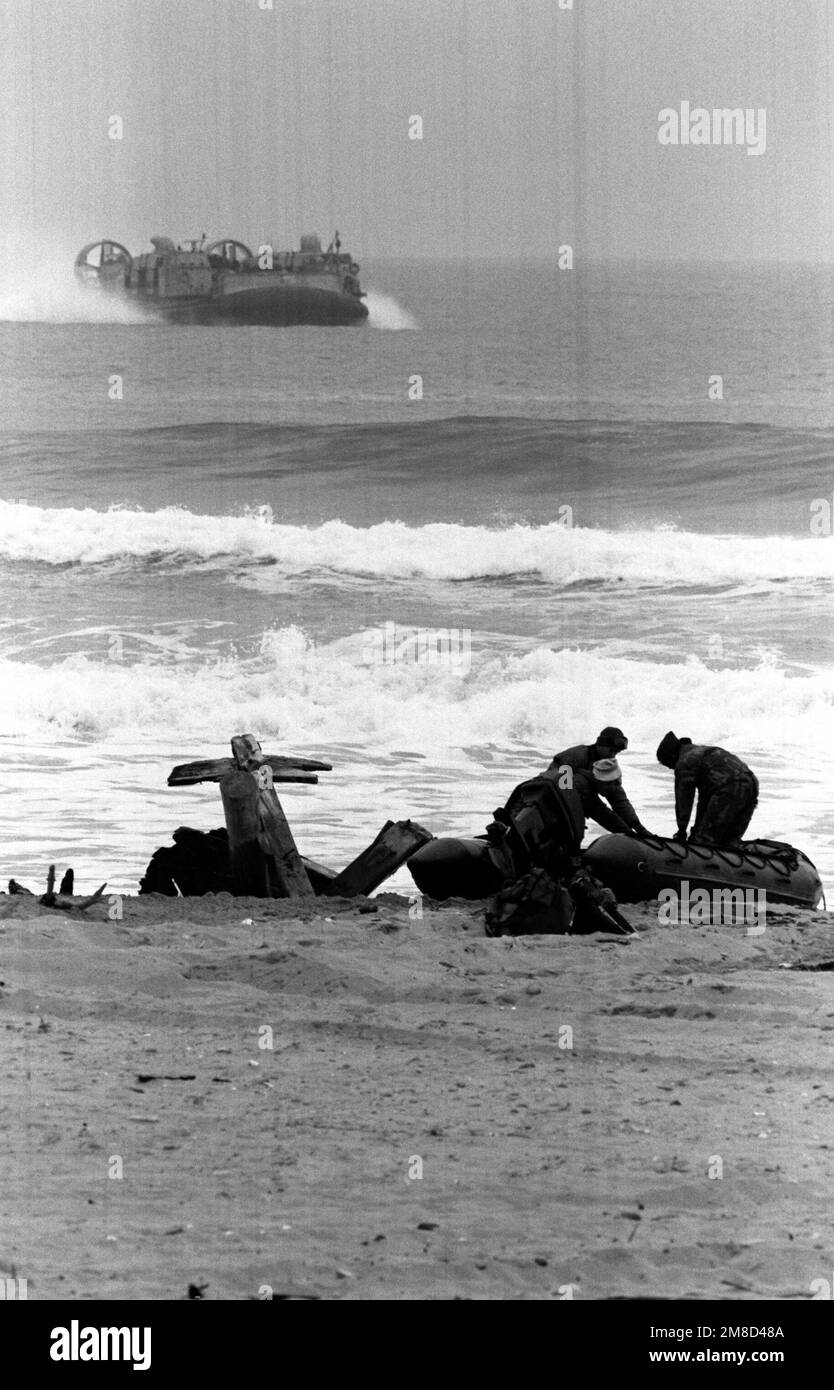 Tre Marines rimuovono la loro attrezzatura da una barca gonfiabile sulla spiaggia come un aereo di atterraggio del cuscino d'aria (LCAC) si avvicina da offshore durante il sud coreano/Stati Uniti che esercitano il Team Spirit '90 combinato. Subject Operation/Series: TEAM SPIRIT '90 base: Pohang Paese: Repubblica di Corea (KOR) Foto Stock