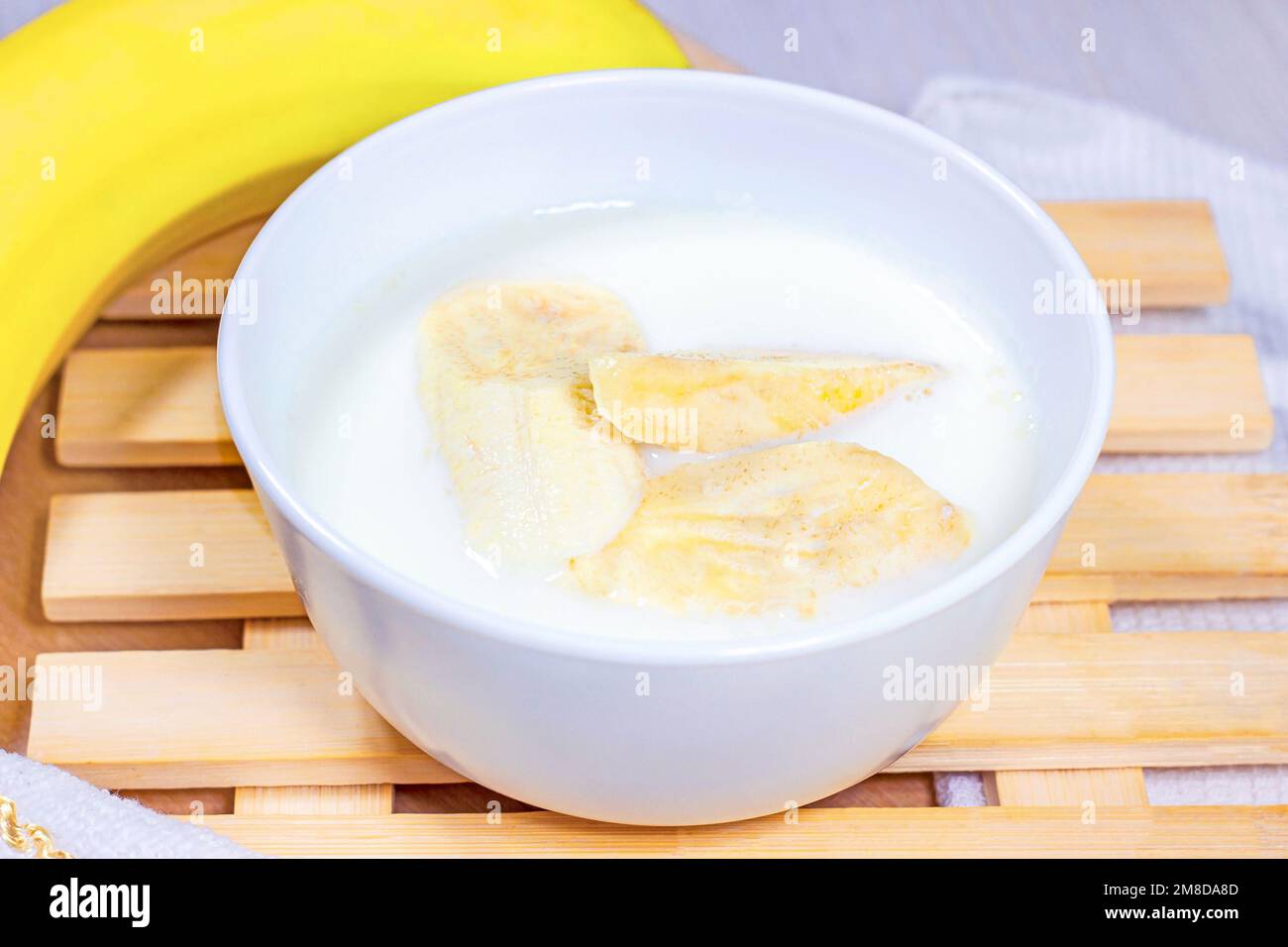 Fette di banana in latte di cocco in una ciotola bianca su sfondo di legno chiaro. Foto Stock