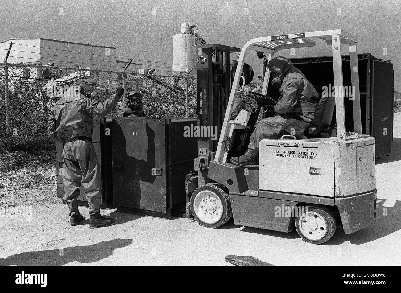 I soldati che indossano un equipaggiamento di protezione completa guidano il conducente di un carrello elevatore a forche mentre abbassa un contenitore sigillato di munizioni chimiche a terra. I membri del 71st Chemical Regiment, 25th Infantry Division (Light), stanno partecipando a un esercizio progettato per testare la capacità dell'unità di rilevare e mitigare una perdita chimica. Base: US Army Chemical Activity Paese: Johnston Atoll Foto Stock