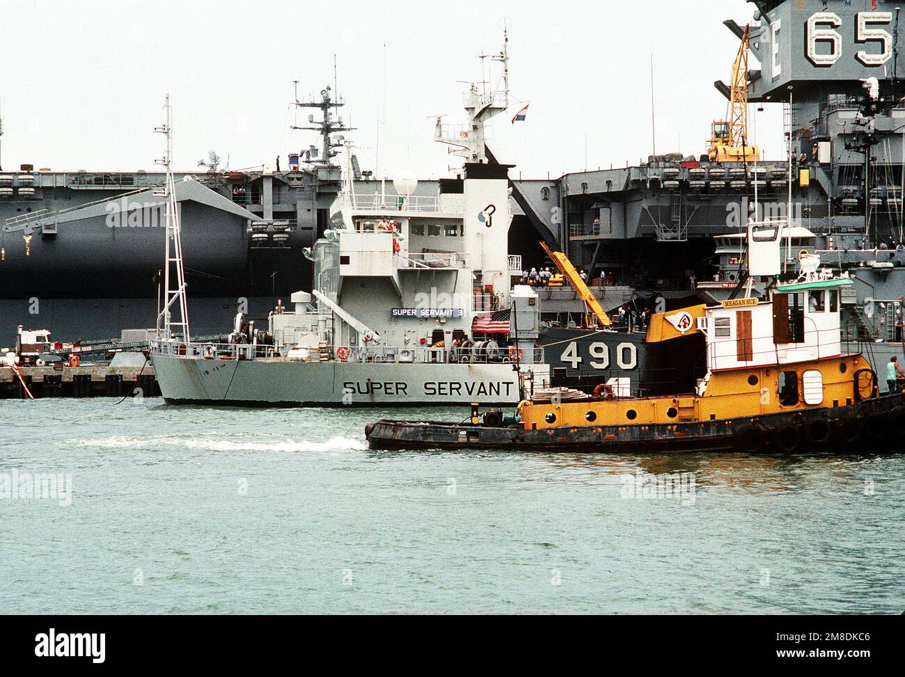 Un rimorchiatore passa oltre la sovrastruttura parzialmente sommersa della nave pesante olandese SUPER SERVER 3. Tre Stati Uniti I minesweepers dell'oceano della Marina e una nave di contromisure della miniera stanno caricando a bordo del SUPER SERVITORE 3 per il trasporto al golfo persiano, dove sosterranno le forze degli Stati Uniti inviate nella regione in reazione all'invasione dell'Iraq del Kuwait. La portaerei a energia nucleare USS ENTERPRISE (CVN-65) è in secondo piano. Soggetto operativo/Serie: DESERT SHIELD/STORM base: Naval Air Station, Norfolk Stato: Virginia (VA) Paese: Stati Uniti d'America (USA) Foto Stock