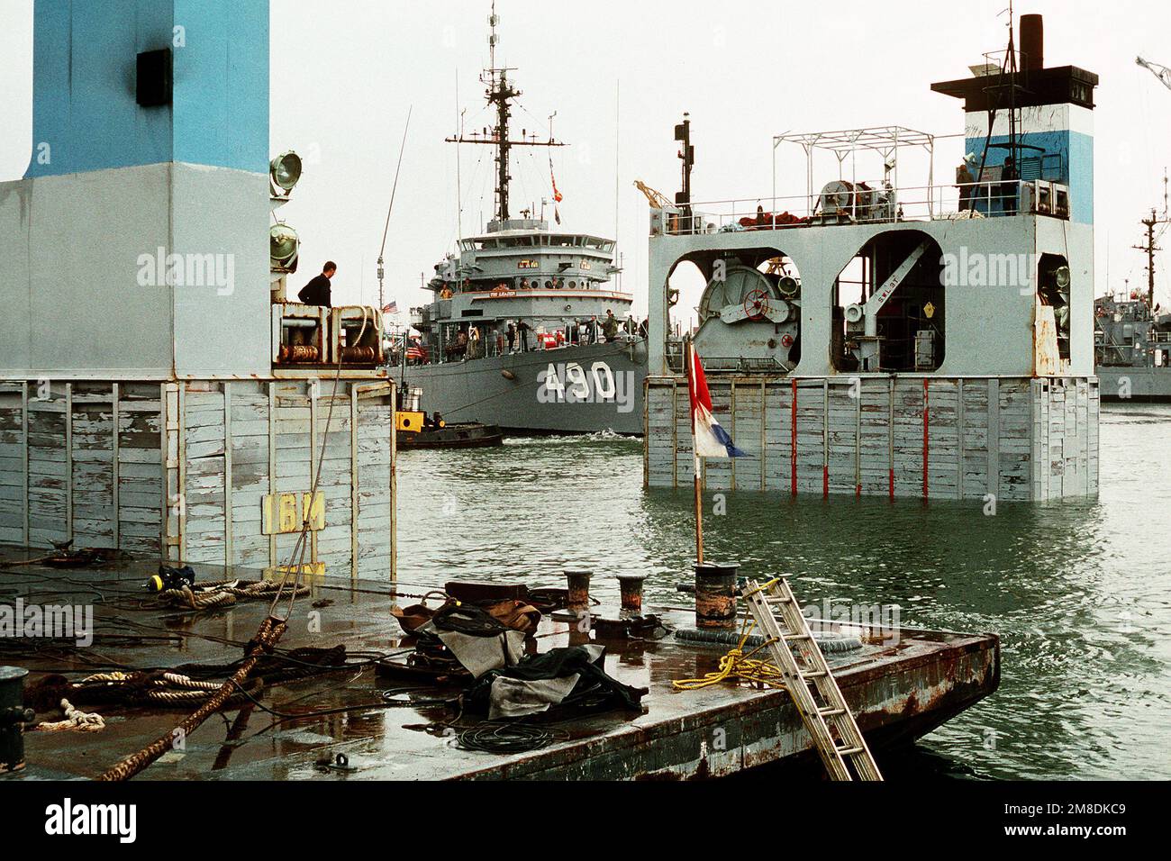Una barca a rimorchiatore tira il LEADER USS (MSO-490) sul ponte sommerso del ponte pesante olandese sh SUPER SERVER 3. Tre Stati Uniti I minesweepers dell'oceano della Marina e una nave di contromisure della miniera stanno caricando a bordo del SUPER SERVITORE 3 per il trasporto al golfo persiano, dove sosterranno le forze degli Stati Uniti inviate nella regione in reazione all'invasione dell'Iraq del Kuwait. La portaerei a energia nucleare USS ENTERPRISE (CVN-65) è in secondo piano. Soggetto operativo/Serie: DESERT SHIELD/STORM base: Naval Air Station, Norfolk Stato: Virginia (VA) Paese: Stati Uniti d'America (USA) Foto Stock