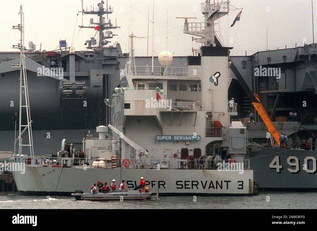 I lavoratori di una piccola imbarcazione passano accanto alla sovrastruttura parzialmente sommersa della nave pesante olandese SUPER SERVER 3. Tre Stati Uniti I minesweepers dell'oceano della Marina e una nave di contromisure della miniera stanno caricando a bordo del SUPER SERVITORE 3 per il trasporto al golfo persiano, dove le forze degli Stati Uniti stanno operando in risposta all'invasione dell'Iraq del Kuwait. La portaerei a energia nucleare USS ENTERPRISE (CVN-65) è in secondo piano. Base: Naval Air Station, Norfolk Stato: Virginia (VA) Paese: Stati Uniti d'America (USA) Foto Stock