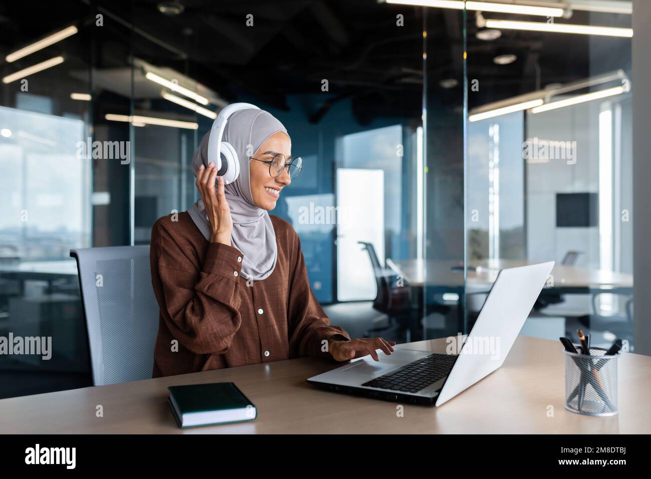 Donna d'affari moderna di successo che lavora all'interno dell'ufficio con un computer portatile, una donna musulmana che indossa un hijab e cuffie che ascolta audiolibri e podcast sul posto di lavoro, donna soddisfatta e di successo. Foto Stock