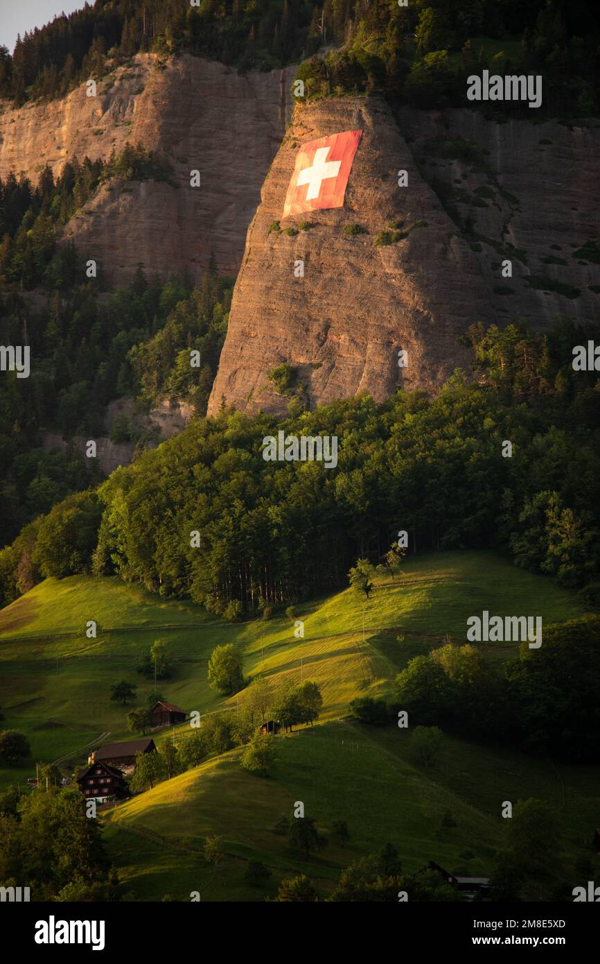 Una montagna svizzera, da lontano, con un piccolo villaggio. sulla roccia appende la bandiera svizzera Foto Stock