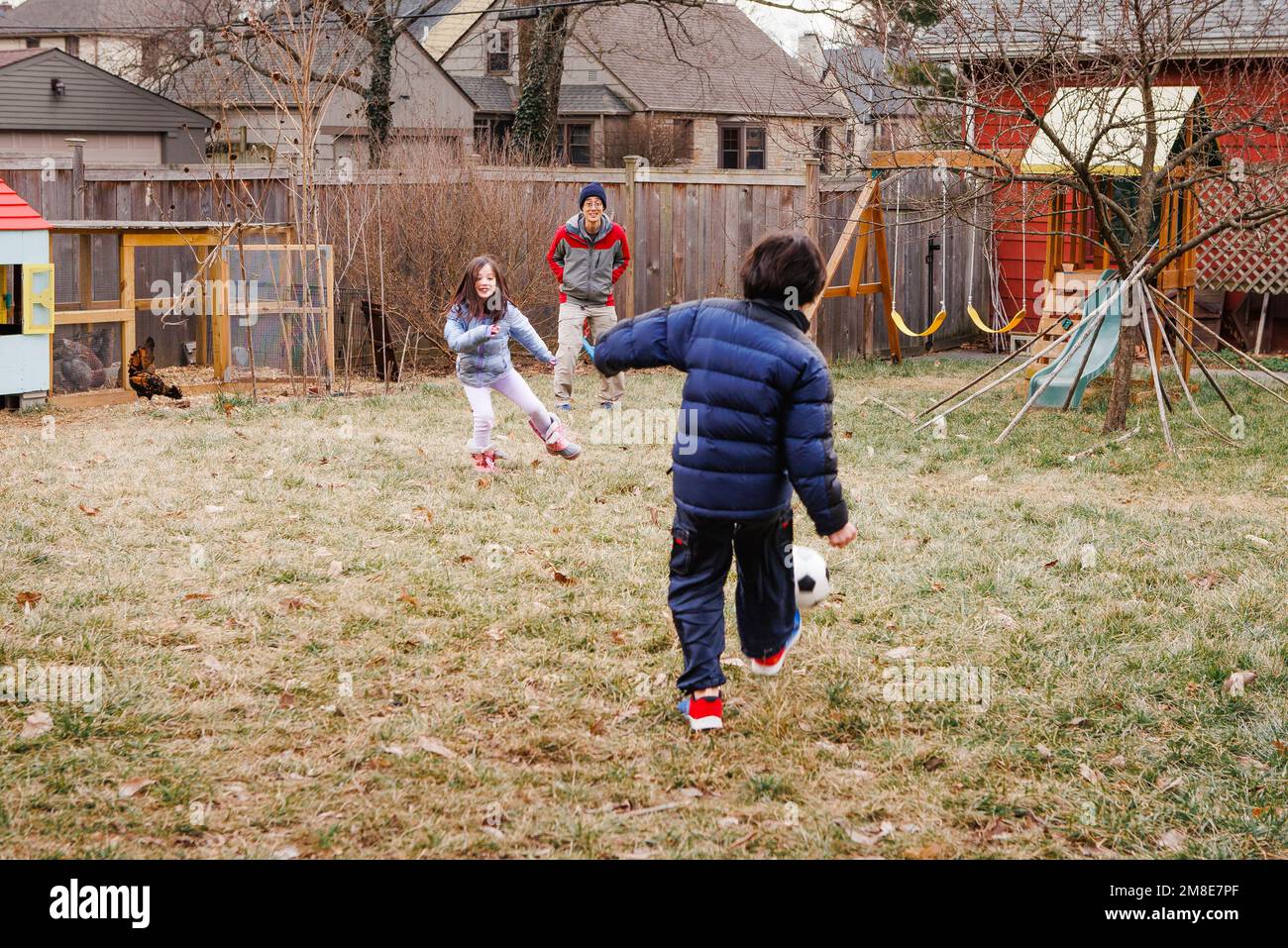 Due bambini giocano a calcio con padre in cortile con polli Foto Stock