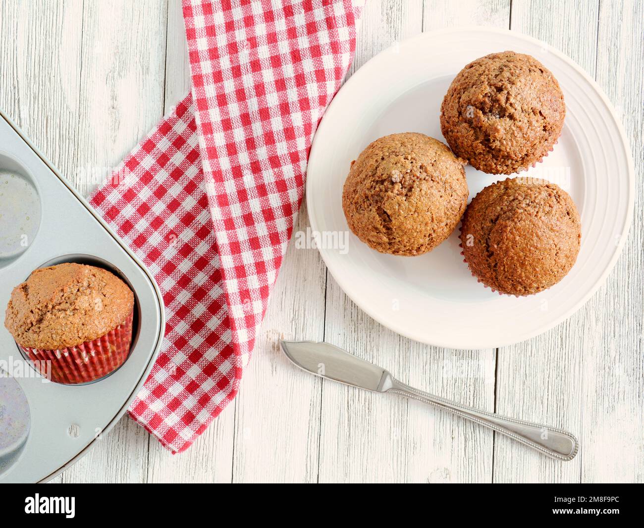 Muffin alla crusca appena sfornati su piatto bianco con tovagliolo rosso a tisana e tovagliolo per muffin su legno bianco rustico in composizione piatta. Foto Stock