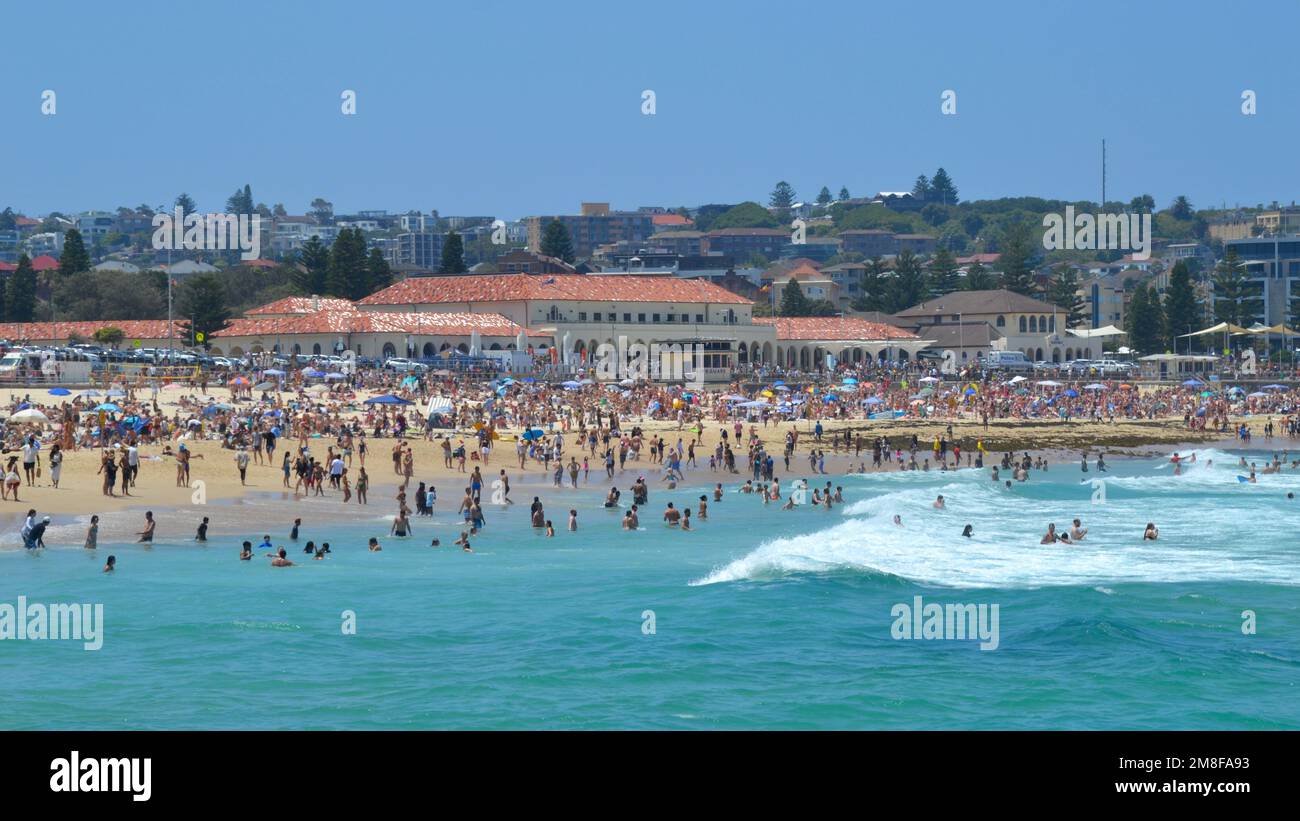 Onde e surf si schiantano sulla sabbia in una trafficata spiaggia di Bondi vicino a Sydney in una perfetta giornata estiva con l'iconico padiglione balneare sullo sfondo Foto Stock