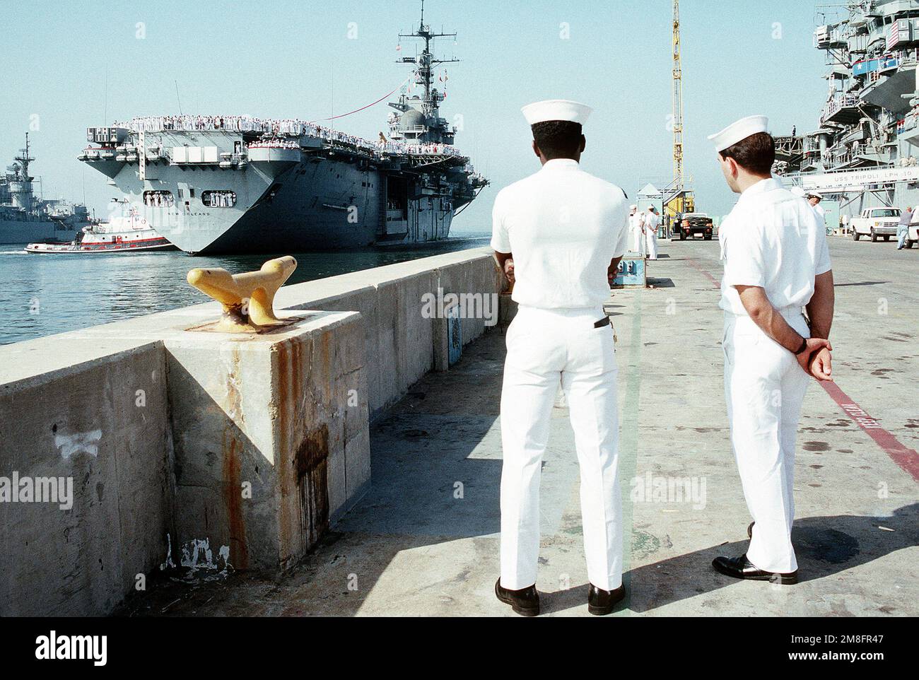 I marinai guardano dal molo mentre un grande rimorchiatore del porto manovra la nave d'assalto anfibio USS NEW ORLEANS (LPH-11) nel porto. La NUOVA ORLEANS, insieme ad altre navi del comandante, lo Squadrone anfibio 1, sta ritornando dall'area del Golfo Persico dove è stata dispiegata durante l'operazione Desert Storm. Soggetto operativo/Serie: DESERT STORM base: Naval Air Station, San Diego Stato: California(CA) Paese: Stati Uniti d'America (USA) Foto Stock