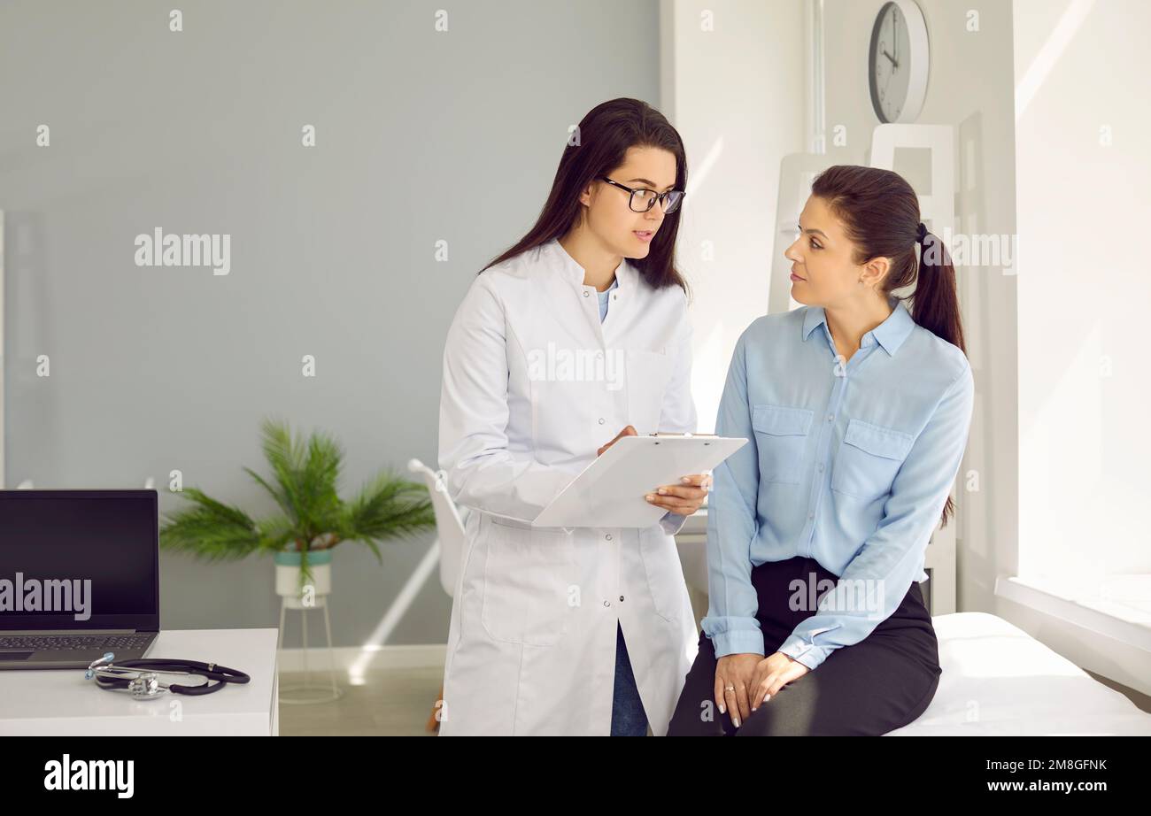 Giovane donna medico in cappotto medico bianco consulta paziente femminile siede sul divano dell'ospedale Foto Stock