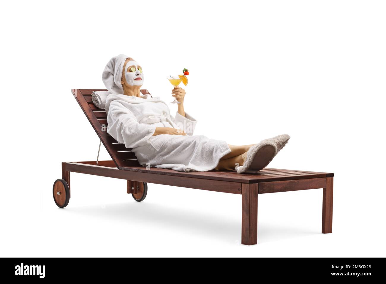 Donna in un accappatoio posata con una maschera viso e cetrioli sugli occhi e tenendo un bicchiere di cocktail isolato su sfondo bianco Foto Stock