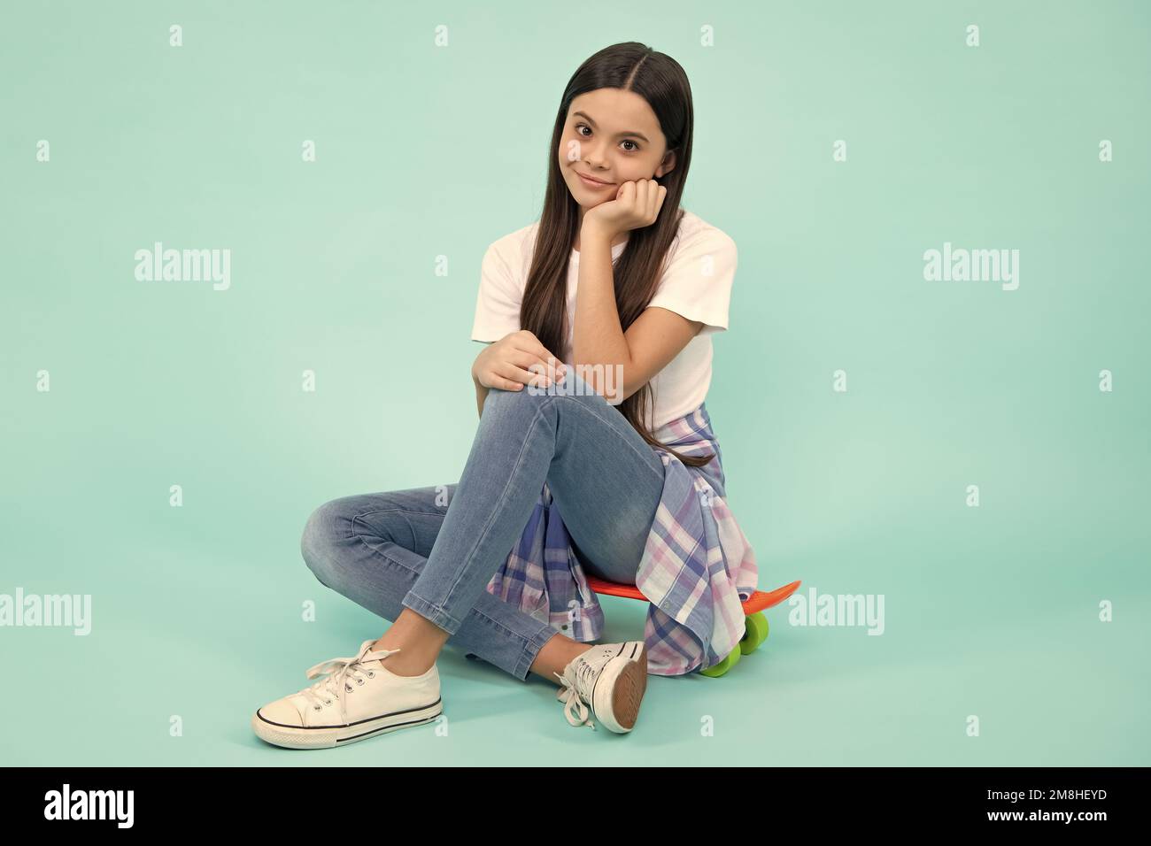 Una ragazza alla moda che indossa una t-shirt alla moda e jeans si siede sullo skateboard, posando su sfondo blu. Moda adolescente, look casual. Felice adolescente, positivo Foto Stock