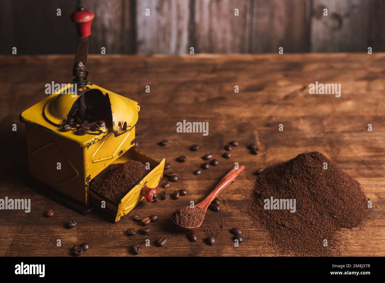 Macinacaffè accanto a un cucchiaio con caffè, una piccola montagna di caffè macinato e chicchi di caffè sparsi sul tavolo di legno Foto Stock