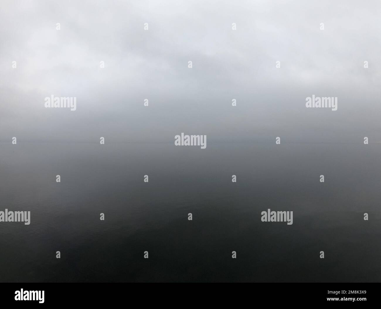 Mare calmo clima nuvoloso. Superficie del mare liscia e senza onde in condizioni di tempo nuvoloso senza vento. Calma. Nuvole di pioggia grigie, linea dell'orizzonte, superficie piana del mare. Tempo piovoso. Grigio bianco astratto sfondo naturale. Foto Stock