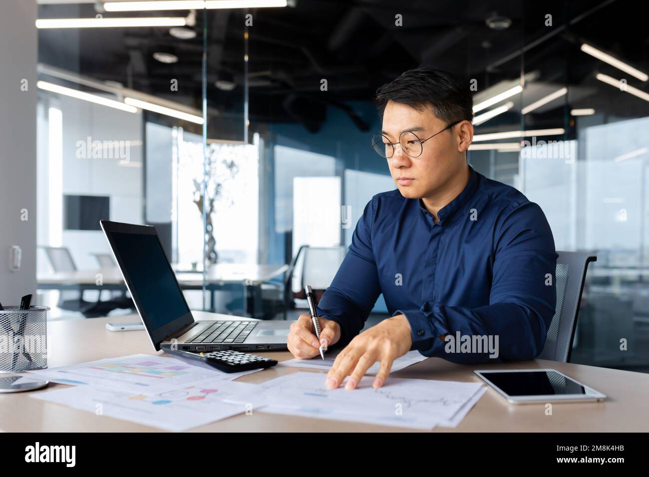 Concentrato giovane uomo d'affari asiatico lavora in ufficio al tavolo con computer portatile, scrive documenti, grafici, accordo, fatture. Foto Stock