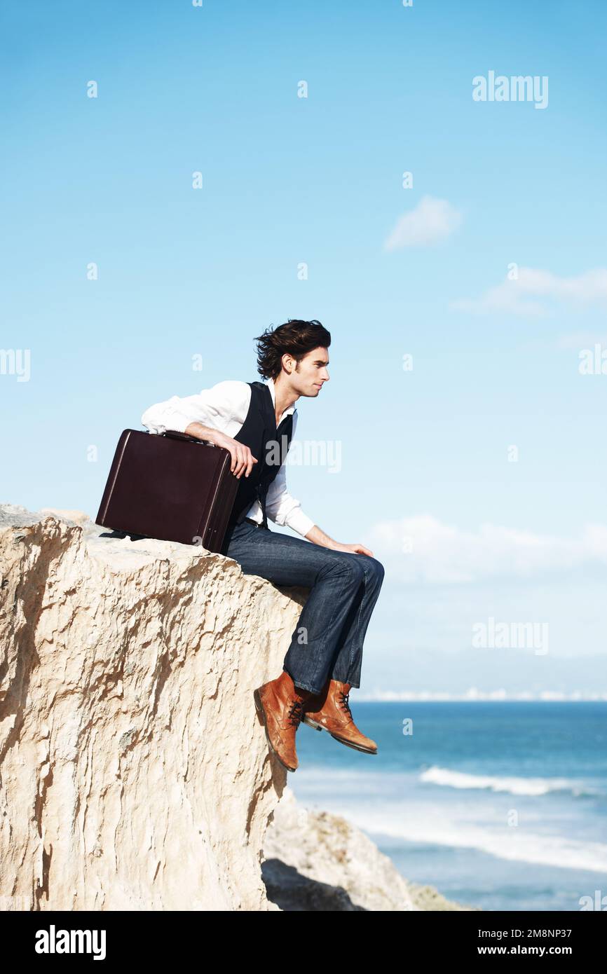 Riflettere sul significato della vita. Giovane uomo pensivo seduto con la sua valigetta sul bordo di una scogliera che domina l'oceano. Foto Stock