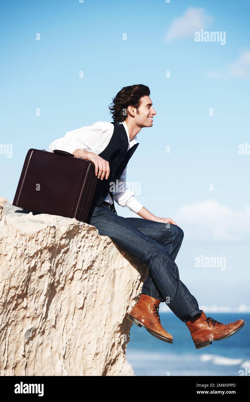 Tornare alle nozioni di base. Giovane uomo sorridente seduto con la sua valigetta sul bordo di una scogliera che domina l'oceano. Foto Stock