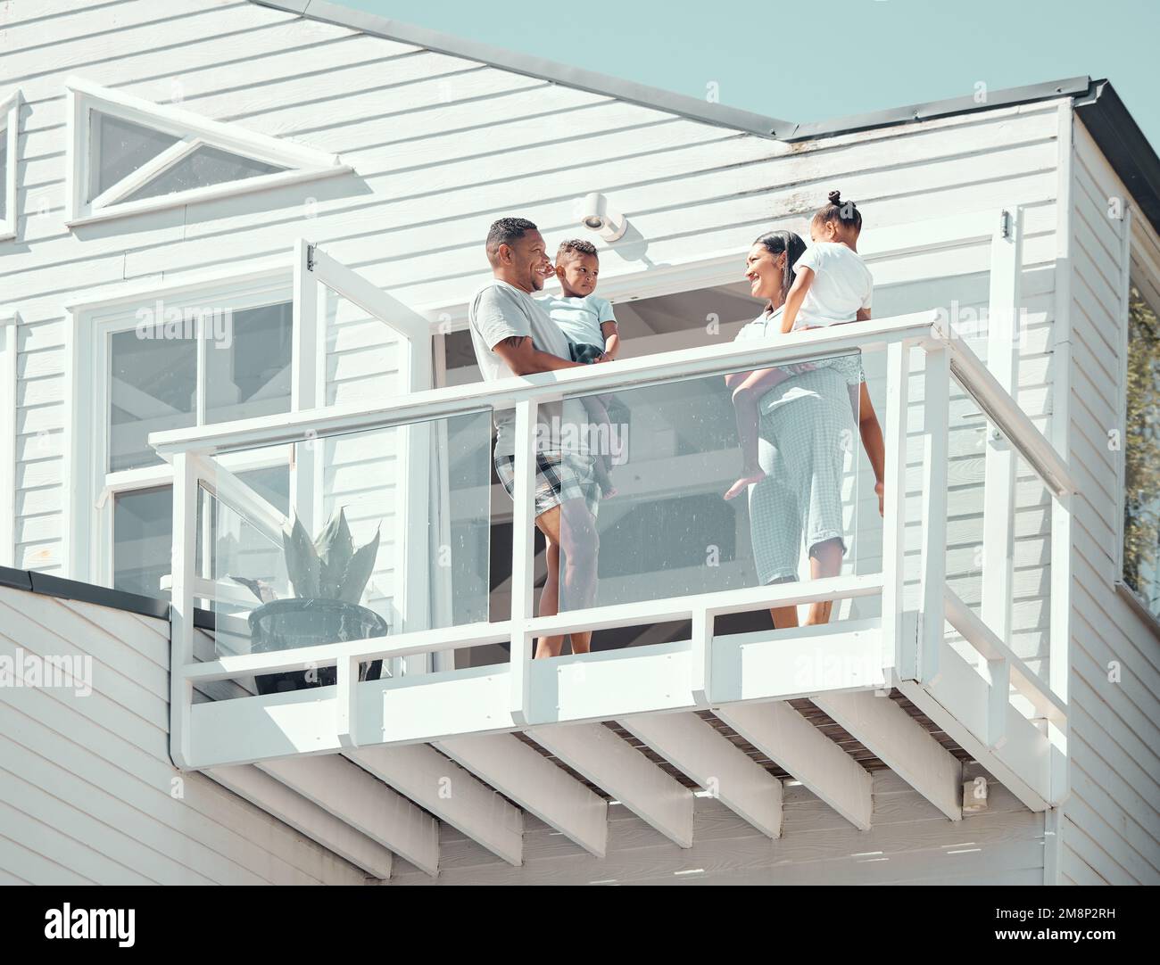 Felice famiglia mista corsa con due bambini che indossano pigiama mentre in piedi sul balcone presso la loro nuova casa o durante le vacanze. Giovani genitori che parlano Foto Stock
