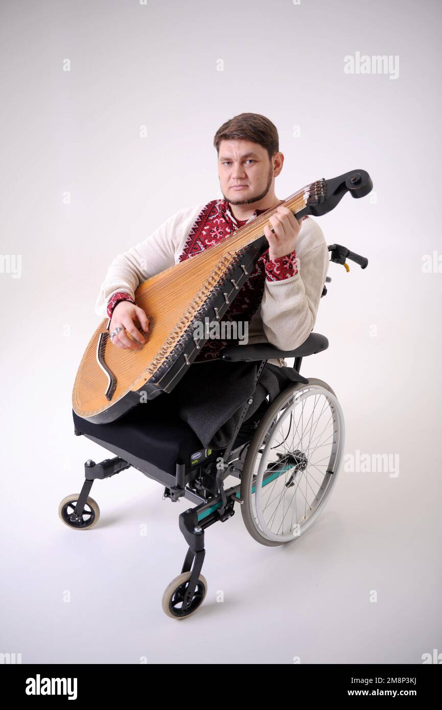 Bel giovane uomo con strumento musicale kobza in mano si siede su sfondo bianco su sedia a rotelle veterano di guerra vestito in camicia Ucraina ricamata na Foto Stock