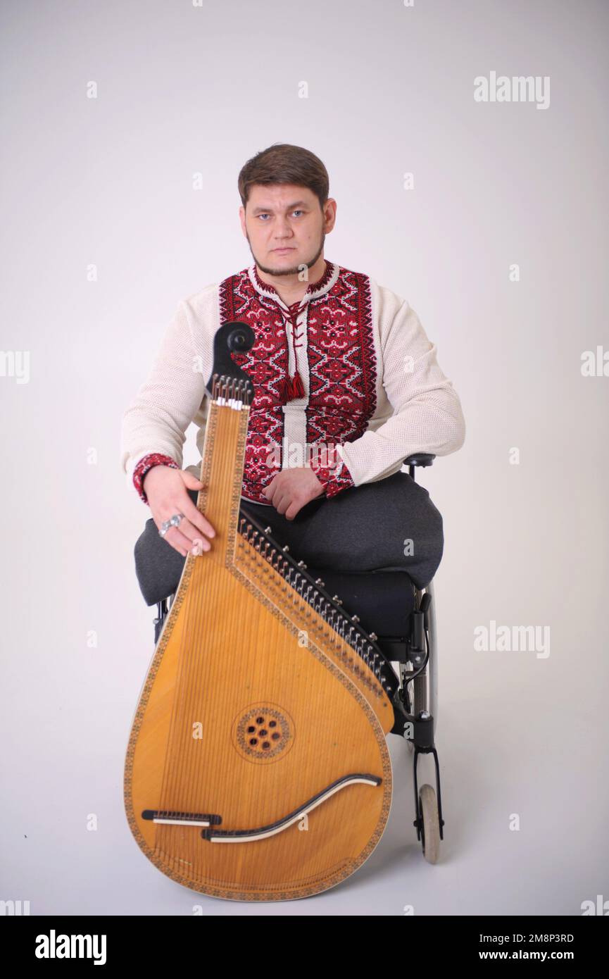 Bel giovane uomo con strumento musicale kobza in mano siede su sfondo bianco veterano di guerra vestito con magliette ucraine ricamati simboli nazionali Foto Stock