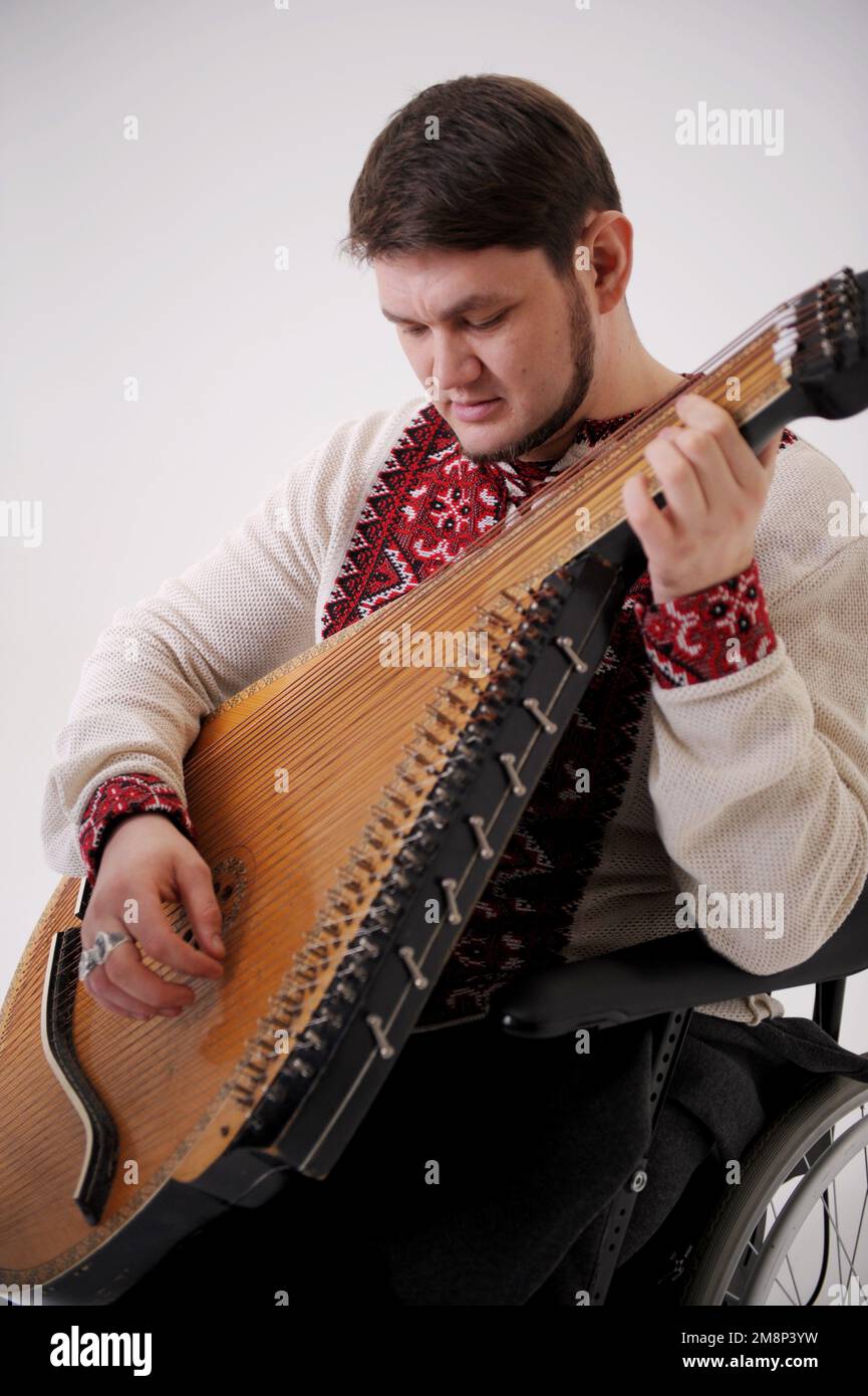 Bel giovane uomo con strumento musicale kobza in mano siede su sfondo bianco veterano di guerra vestito con magliette ucraine ricamati simboli nazionali Foto Stock