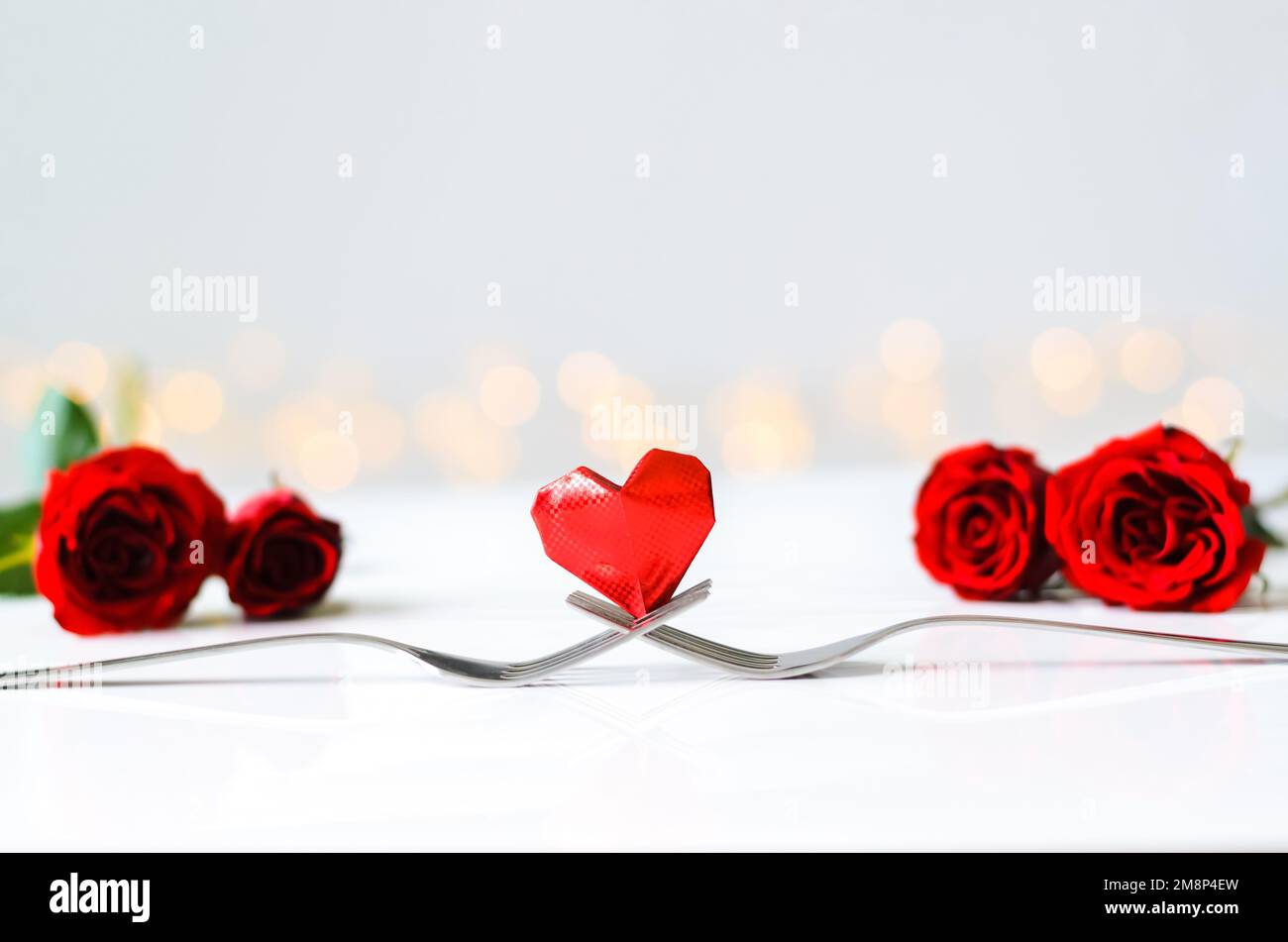 Piega in carta a forma di cuore rosso su due forchette con rose poste su un tavolo bianco e sfondo chiaro bokeh per il concetto di San Valentino. Foto Stock