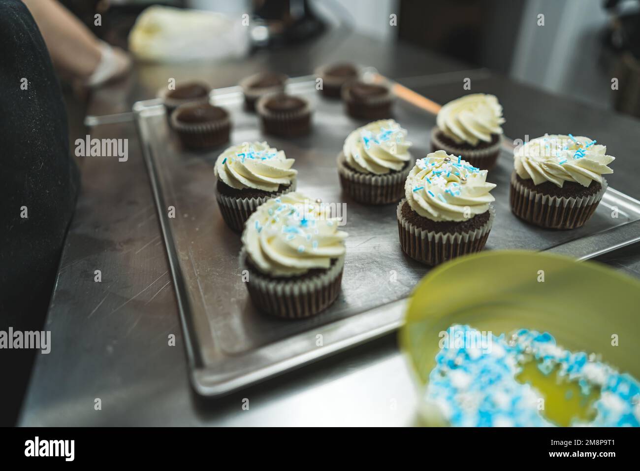 Cupcake al cioccolato con glassa alla vaniglia e spolverini blu e bianchi sul vassoio d'argento Processo professionale di decorazione da forno. Scatto orizzontale in interni. Foto di alta qualità Foto Stock