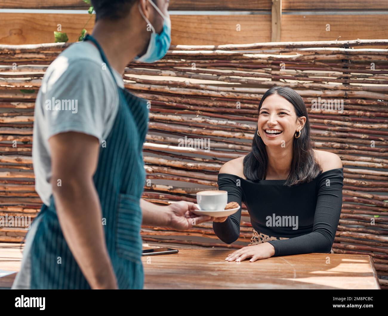Felice giovane donna ispanica che riceve una tazza di caffè da un cameriere che indossa una maschera facciale in un bar locale. Donna di razza mista eccitata per essere servita il suo caldo Foto Stock