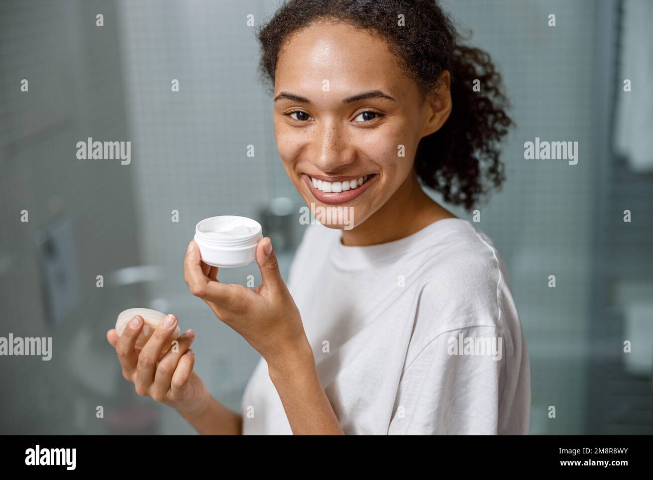 Donna africana sorridente applicando crema idratante in piedi nella stanza da bagno domestica. Procedura di bellezza Foto Stock