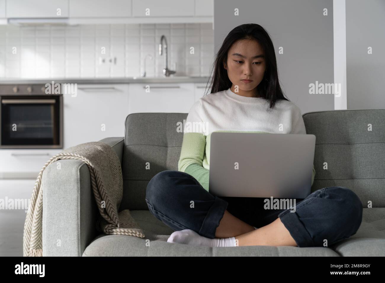 Una donna coreana professionista concentrata siede sul divano con un computer portatile e scrive testo in remoto Foto Stock