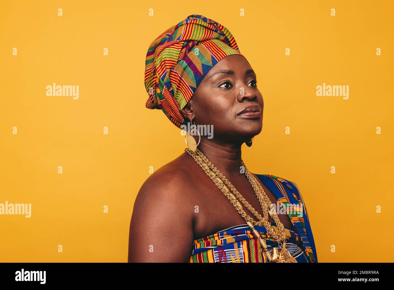 Donna africana matura che indossa abiti tradizionali su sfondo giallo. Donna nera sicura vestita in tessuto colorato Kente e gioiello d'oro Foto Stock