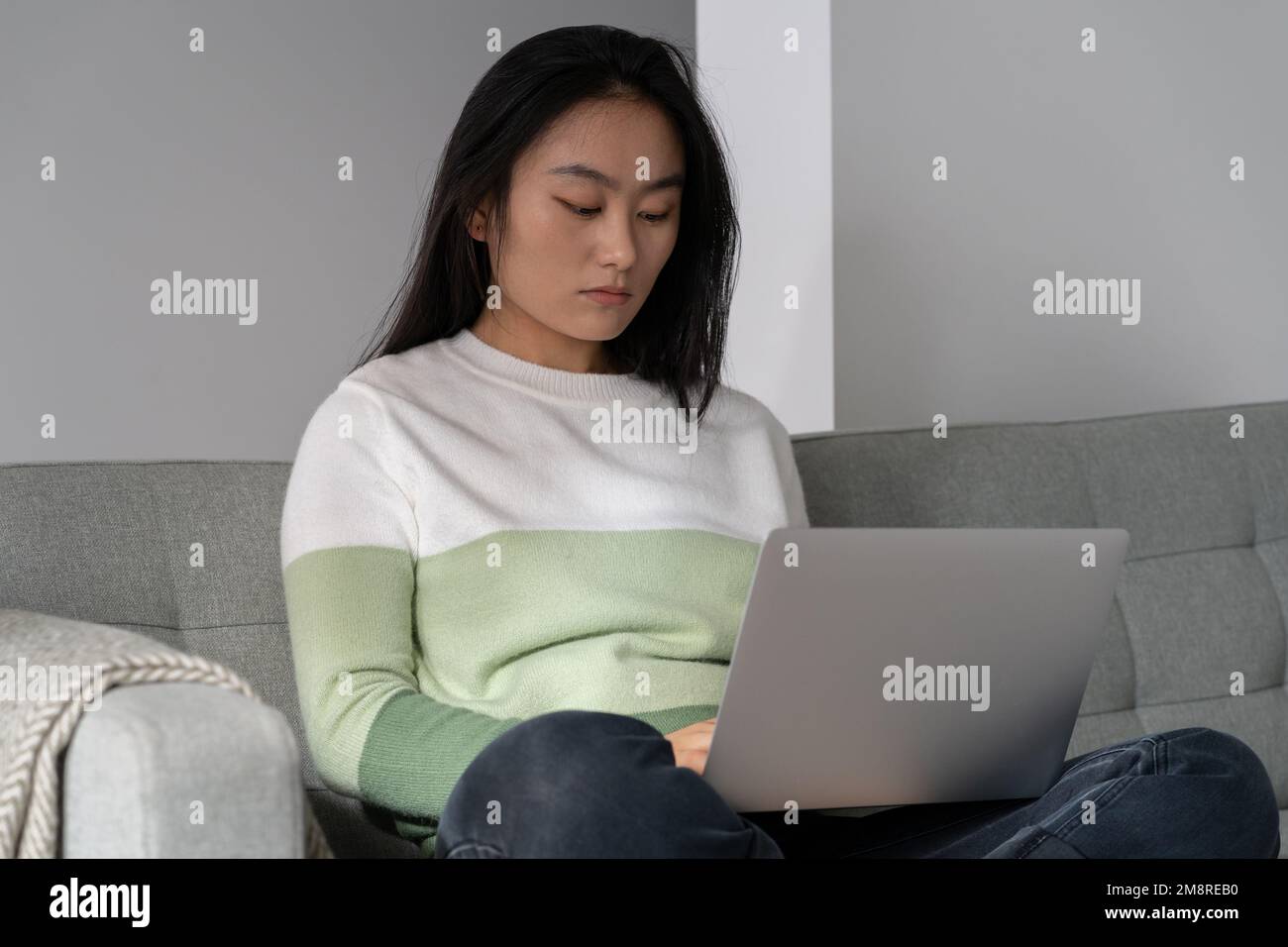 Donna asiatica concentrata freelancer con notebook in grembo si siede sul divano a distanza lavorando da casa Foto Stock